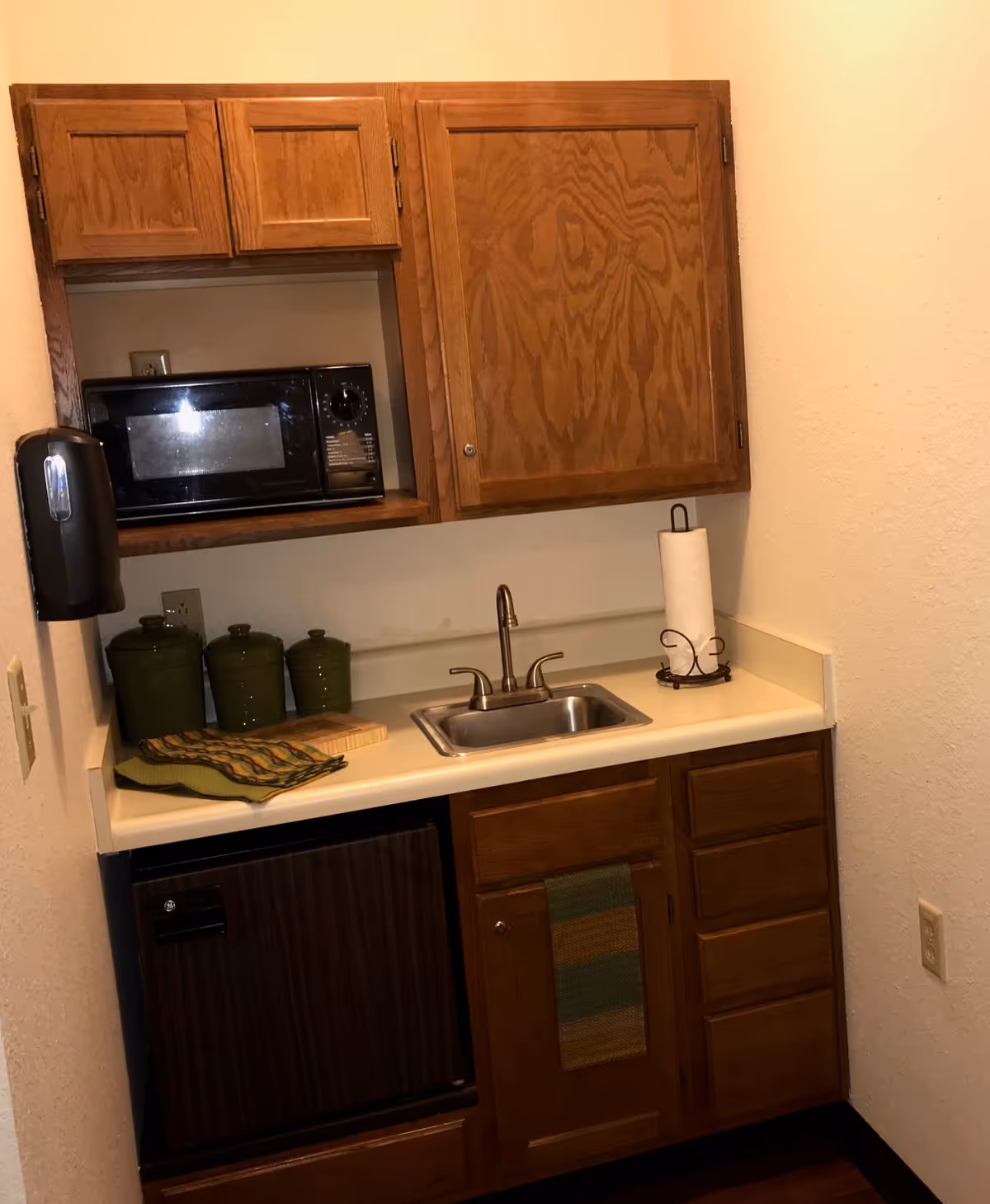 Small kitchenette with wooden cabinets, a microwave, a stainless steel sink with a faucet, a paper towel holder, three green canisters, a cutting board, and a small refrigerator below the counter.