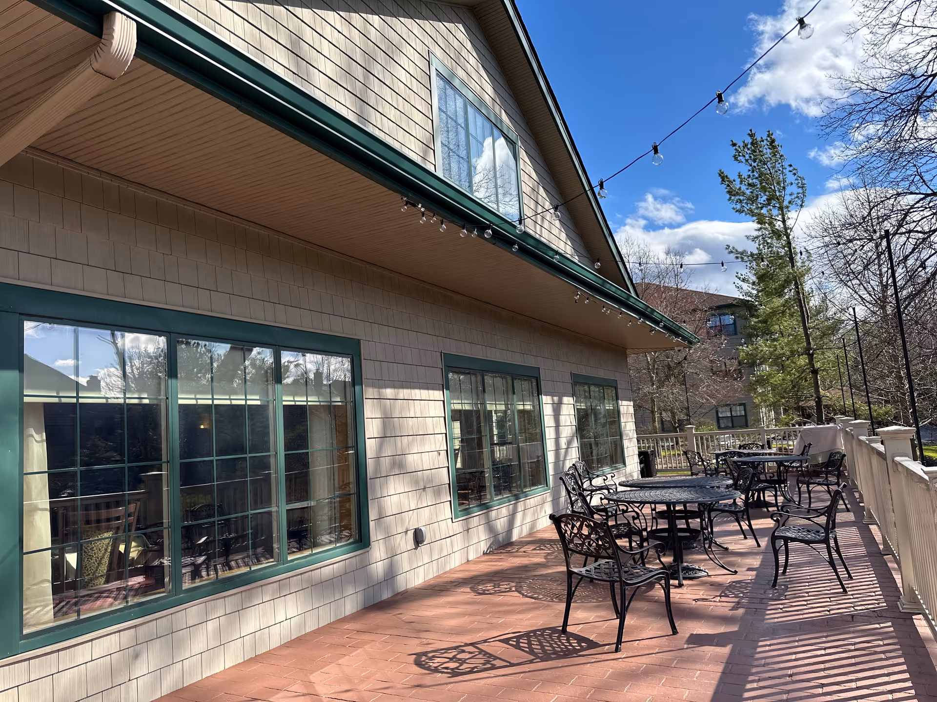 Outdoor patio area at Deer Creek Lodge with several black metal tables and chairs on a brick floor. The building has beige siding with green-trimmed windows. String lights are hung above the patio, and trees and a blue sky with clouds are visible in the background.