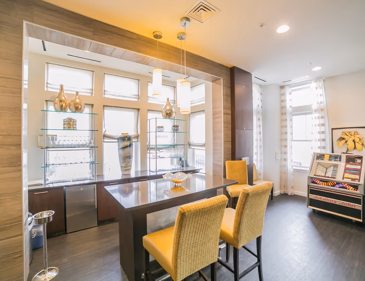 A bright and modern interior room with a high table and four yellow cushioned chairs. The room features large windows with sheer curtains, glass shelves holding decorative vases and glassware, and three pendant lights hanging above the table. There is a vintage jukebox labeled 'Jukebox John' near the windows and a framed artwork on the wall.