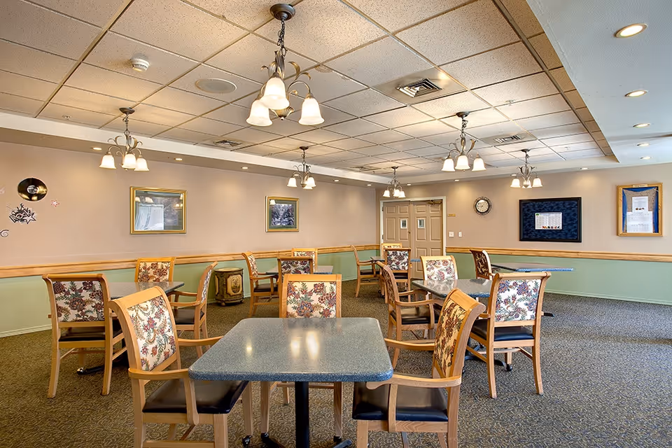 A dining room with multiple tables and chairs arranged neatly. The chairs have wooden frames with floral patterned upholstery. The room has a carpeted floor, beige walls with green wainscoting, and several framed pictures and bulletin boards on the walls. Ceiling lights with multiple bulbs hang from the ceiling, illuminating the space.