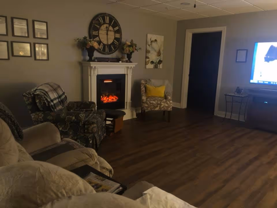 A cozy living room with a beige recliner, a patterned armchair with a plaid blanket, and another floral armchair with a yellow pillow. There is a white electric fireplace with a large decorative clock and flower arrangements on the mantel. The room has wood flooring, framed certificates on the wall, and a TV on a stand in the corner.