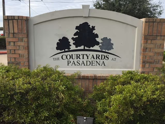 A large sign made of brick and concrete with three tree silhouettes and the text 'The Courtyards at Pasadena' surrounded by green bushes.