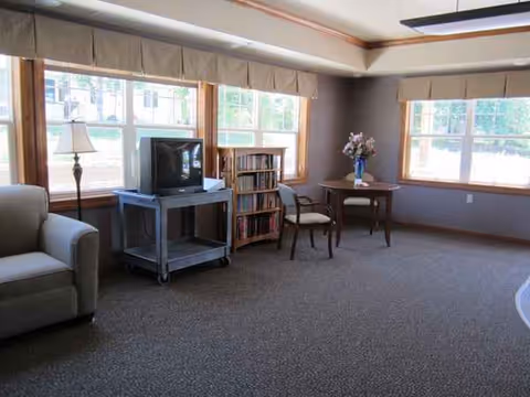A cozy living room in a care facility with sofa, TV cart, bookshelves, and windows.
