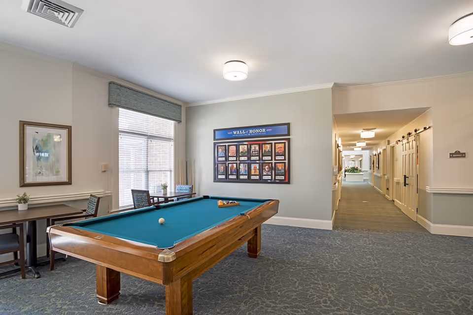 A well-lit recreational room featuring a pool table with balls set up for a game. To the left, there are tables and chairs near a window with blinds and a valance. On the wall above the pool table is a 'Wall of Honor' display with framed photos. A hallway extends to the right with doors and light fixtures along the ceiling.