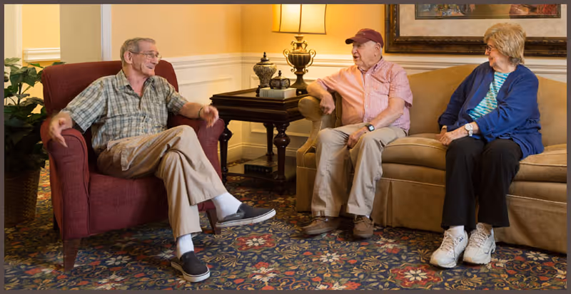 Three elderly people sitting and conversing in a cozy living room area. One man is seated in a red armchair on the left, while another man and a woman sit together on a beige couch. There is a side table with a lamp and decorative items between them, and a patterned carpet on the floor.