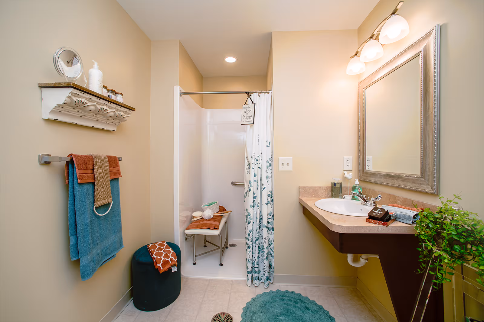 Well-lit bathroom with a shower and curtain, wall-mounted sink and mirror, towels on a rack, and decorative shelf and plant.