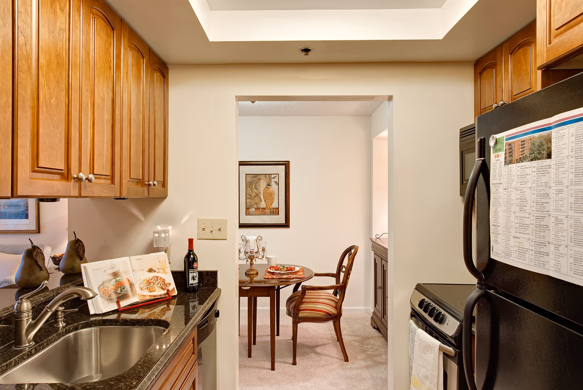 A kitchen area with wooden cabinets, a stainless steel sink, a black granite countertop, and a black refrigerator with a paper attached to it. Beyond the kitchen, there is a small dining area with a round wooden table, two chairs with striped cushions, a framed artwork on the wall, and a decorative candelabra on the table.