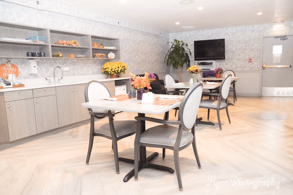 A bright dining room with several tables and chairs arranged neatly. Each table has a small floral centerpiece and place settings. Along one wall, there are cabinets and open shelves decorated with small pumpkins and flowers. A flat-screen TV is mounted on the far wall, and a large potted plant is placed near the corner. The floor has a light wood pattern, and the walls are decorated with a subtle patterned wallpaper.