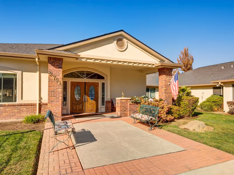 Front entrance of a senior living facility with double wooden doors, brick pillars, two benches with floral cushions, an American flag, and well-maintained landscaping under a clear blue sky.