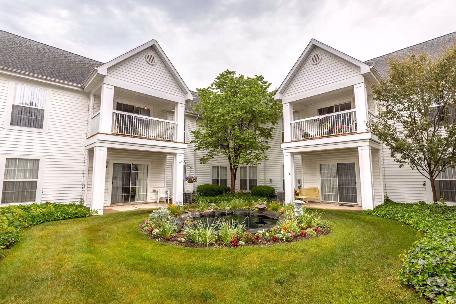 A courtyard area of a senior living facility with a well-maintained green lawn, a circular flower bed with various colorful flowers and plants surrounding a small pond, and two white two-story buildings with balconies and sliding glass doors facing the courtyard. Trees and shrubs are also visible around the buildings.
