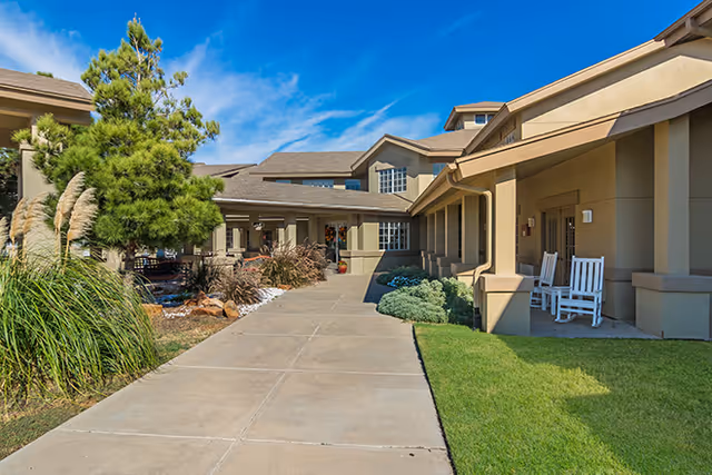 Exterior view of a senior living facility with a wide concrete walkway leading to the entrance. The building has beige walls and a covered porch area with white rocking chairs. There are green shrubs, ornamental grasses, and a tree along the walkway under a clear blue sky.
