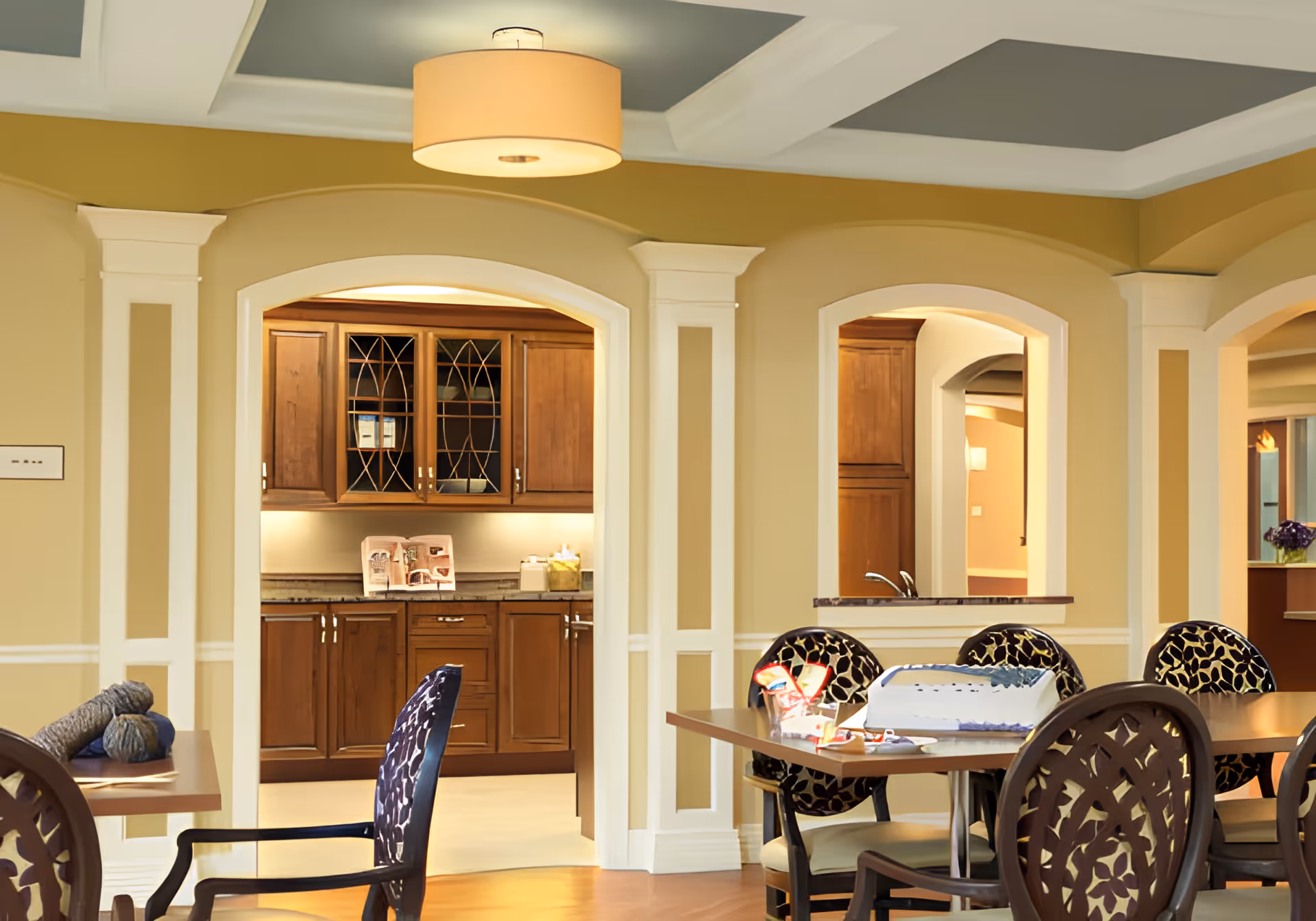 Interior view of a senior living facility dining area with wooden tables and patterned chairs. In the background, there is a kitchen area with wooden cabinets and a countertop. The room features beige walls with white trim and a ceiling light fixture.