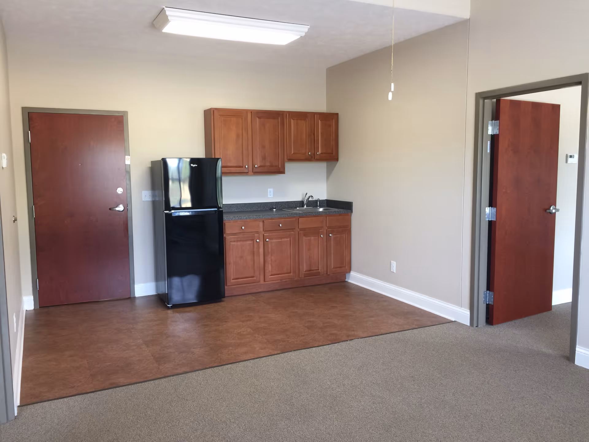 A small kitchen area in a retirement community apartment featuring wooden cabinets, a black refrigerator, a sink with a countertop, and two closed wooden doors. The floor transitions from brown tile in the kitchen area to carpet in the adjacent room.