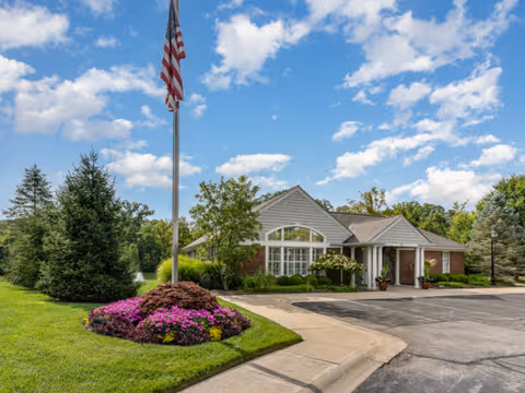 Exterior view of a single-story building with a gabled roof, surrounded by well-maintained landscaping including green grass, trees, and a flower bed with purple flowers. An American flag is flying on a flagpole near the sidewalk under a partly cloudy blue sky.