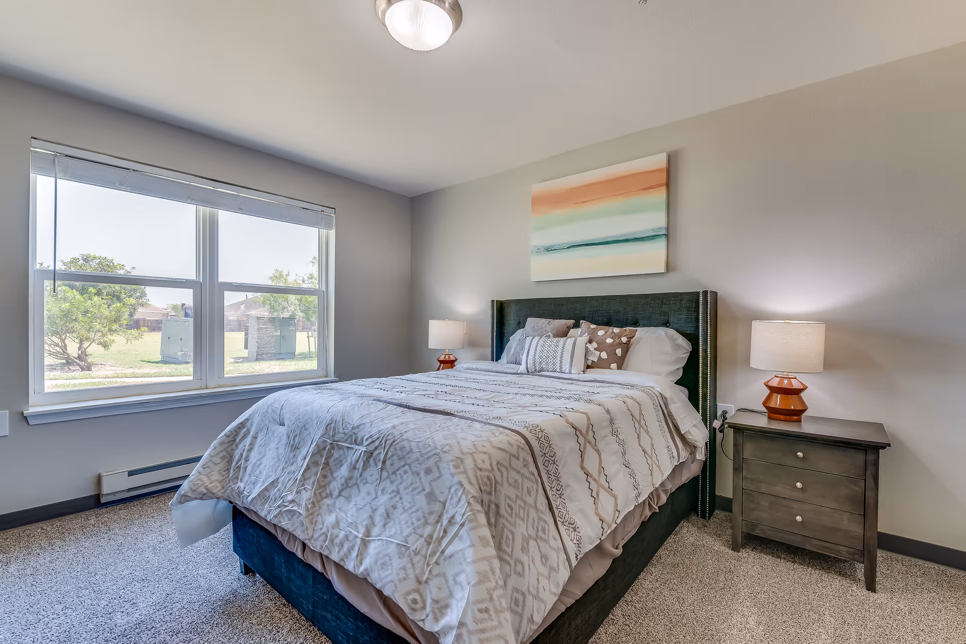 A bright bedroom with a large window showing an outdoor view. The room features a bed with a patterned comforter and multiple pillows, a dark wooden nightstand with a lamp on each side, and a colorful abstract painting hanging above the bed.