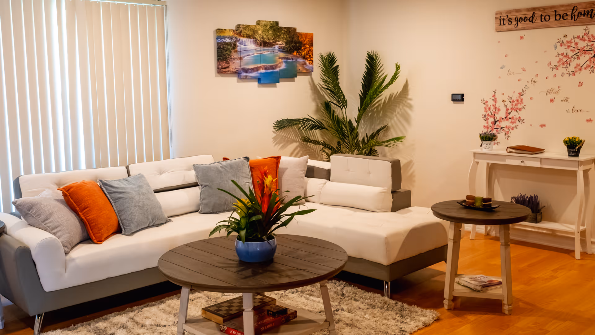 Well-lit living room with a white sectional sofa, colorful throw pillows, round wooden coffee tables, potted plants, and wall decor.