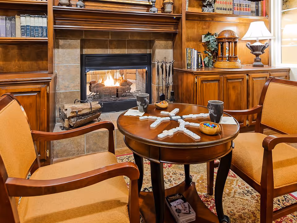 Cozy living room with a lit fireplace, a round table arranged with game tiles, mugs and snacks, and upholstered chairs.