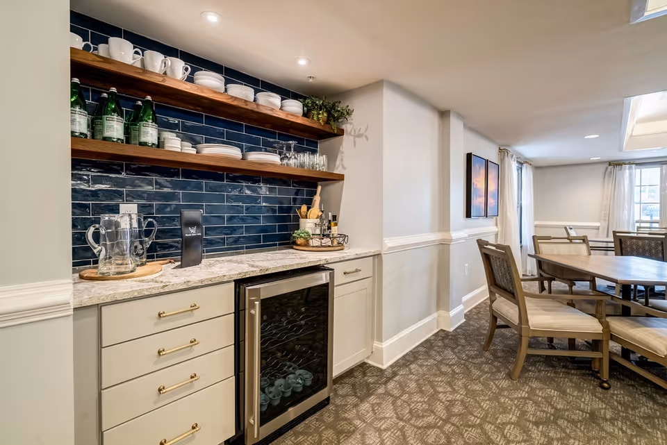 Dining area with round wooden tables and upholstered chairs next to a kitchenette featuring open wooden shelves, a wine cooler, and a blue tile backsplash.