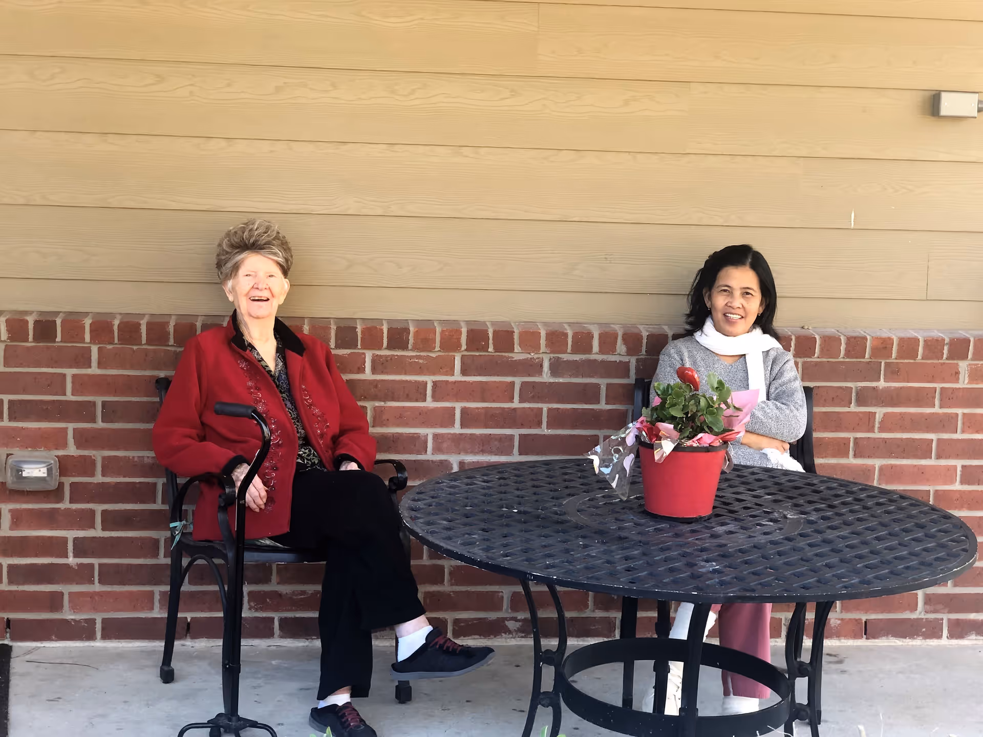 Two women sitting outside on black metal chairs at a round black metal table with a red potted plant on it, against a background of a brick wall and beige siding.