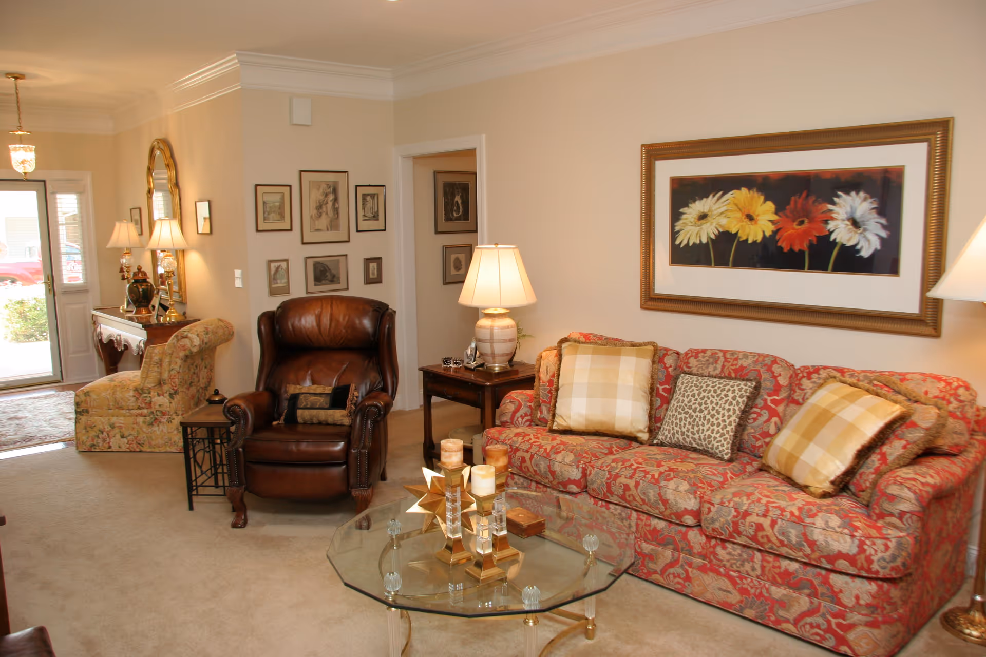 A cozy living room with a floral patterned sofa adorned with three pillows, a brown leather armchair, and a floral upholstered chair near the entrance. The room features a glass coffee table with decorative candles, a side table with a lamp, framed artwork on the walls, and a large framed picture of colorful flowers above the sofa. The space is warmly lit with table lamps and natural light coming through the glass front door.