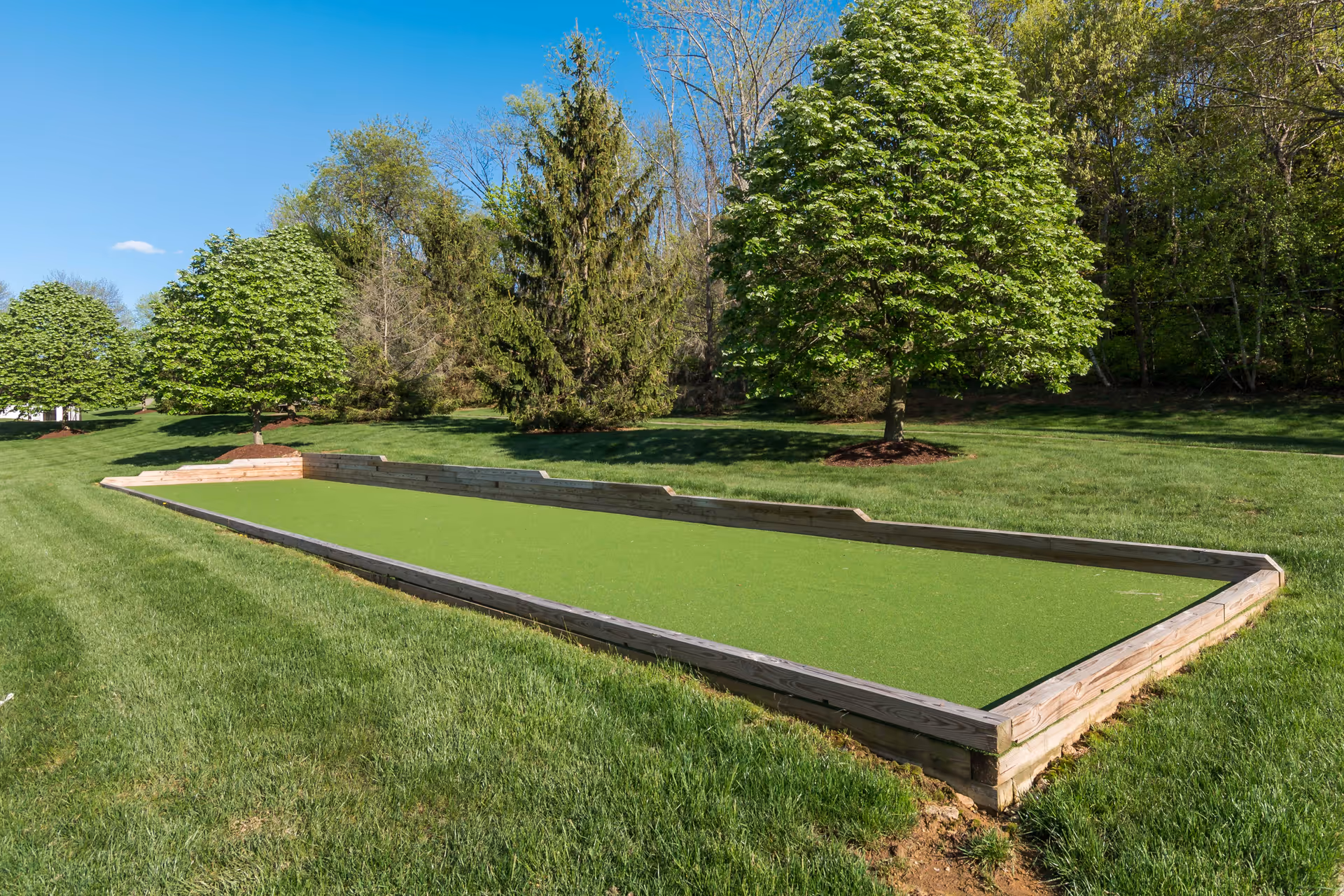 Outdoor bocce ball court surrounded by green grass and trees under a clear blue sky.