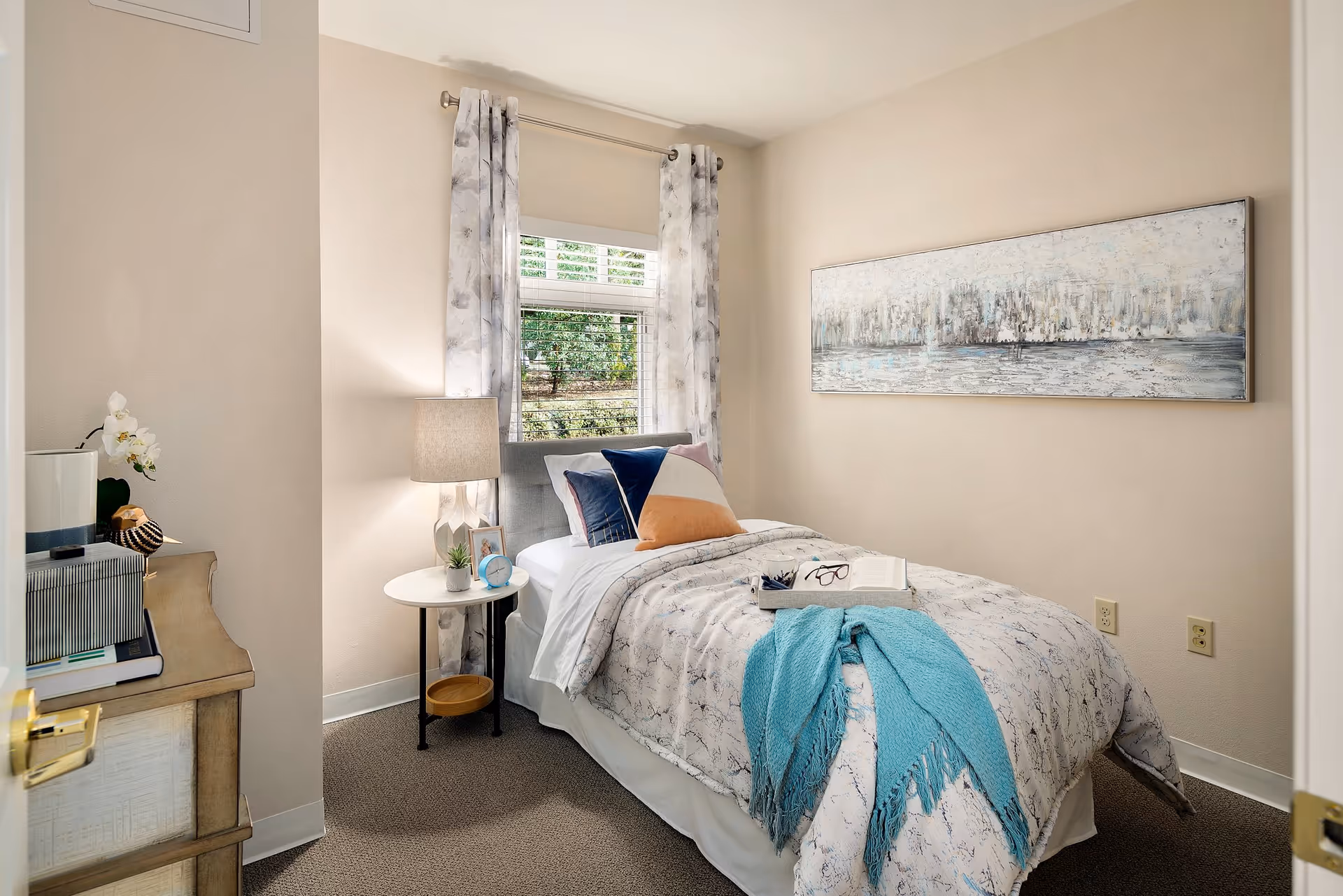 A cozy bedroom with a single bed covered in a patterned comforter and a teal throw blanket. The bed has multiple decorative pillows and a tray with a book and glasses on it. Next to the bed is a round white side table with a lamp, a small plant, a photo frame, and a blue alarm clock. A window with floral curtains lets in natural light, and a large abstract painting hangs on the beige wall.