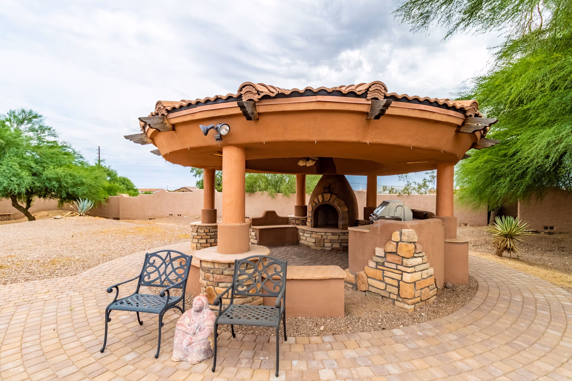 Outdoor covered patio area with a circular roof supported by columns, featuring a built-in stone fireplace, seating benches, a grill, two metal chairs, and a small statue of a seated figure. The area is surrounded by desert landscaping with trees and gravel.