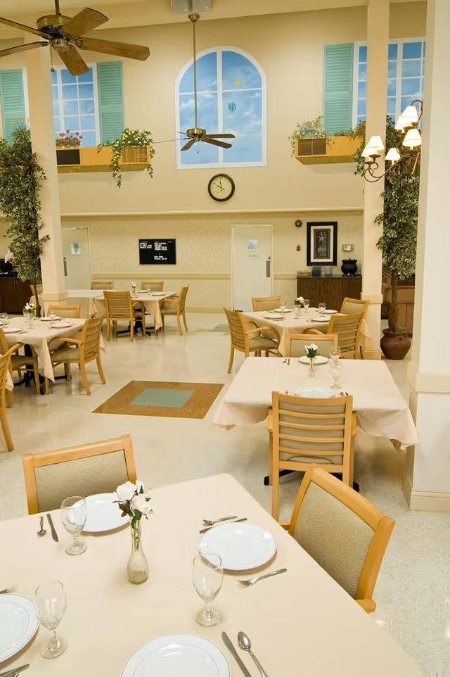 A bright dining room with several tables covered in beige tablecloths, each set with white plates, glasses, and silverware. The room features wooden chairs, ceiling fans, and a wall mural depicting windows with blue shutters and flower boxes. There are plants and wall sconces adding to the decor.