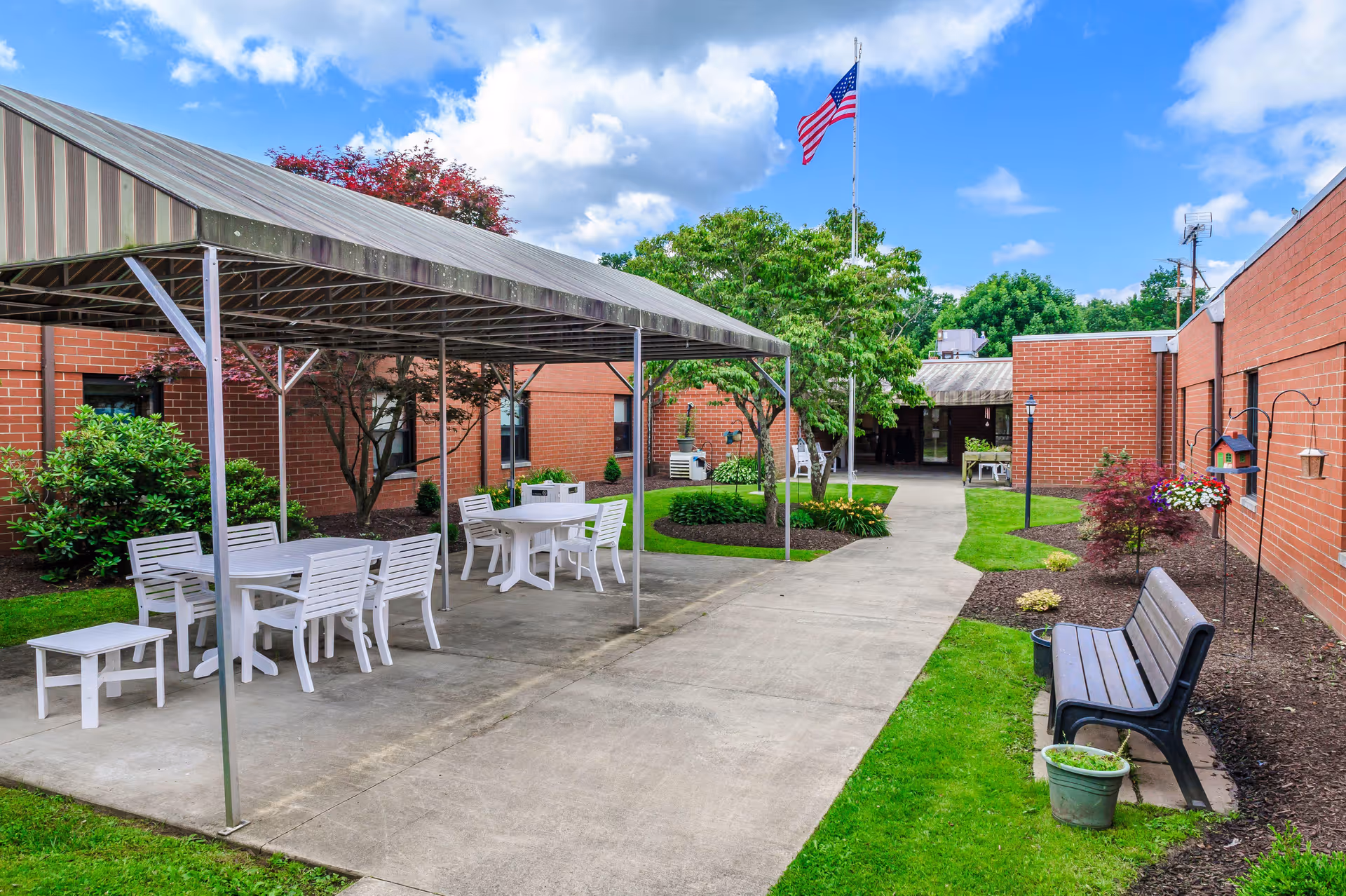 Outdoor courtyard area at Kittanning Health & Rehab Center featuring a covered seating area with white tables and chairs, a concrete walkway, green grass, trees, and an American flag on a flagpole against a blue sky with clouds.