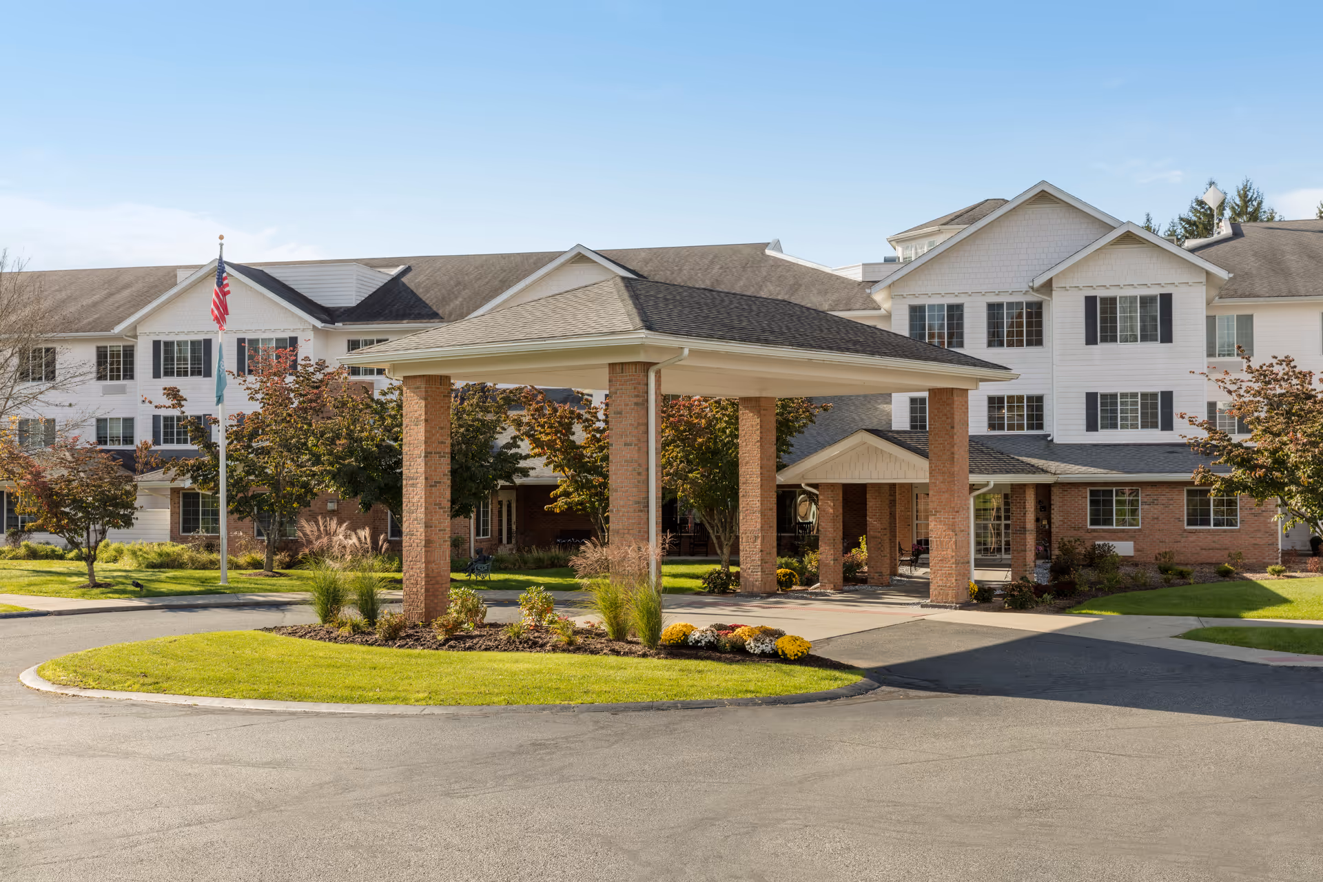 Exterior view of Holiday Bluebird Estates, a multi-story senior living facility with white siding and brick accents. The entrance features a covered drop-off area supported by brick columns, surrounded by well-maintained landscaping including green grass, bushes, and flowers. Two flagpoles with flags are visible near the entrance under a clear blue sky.