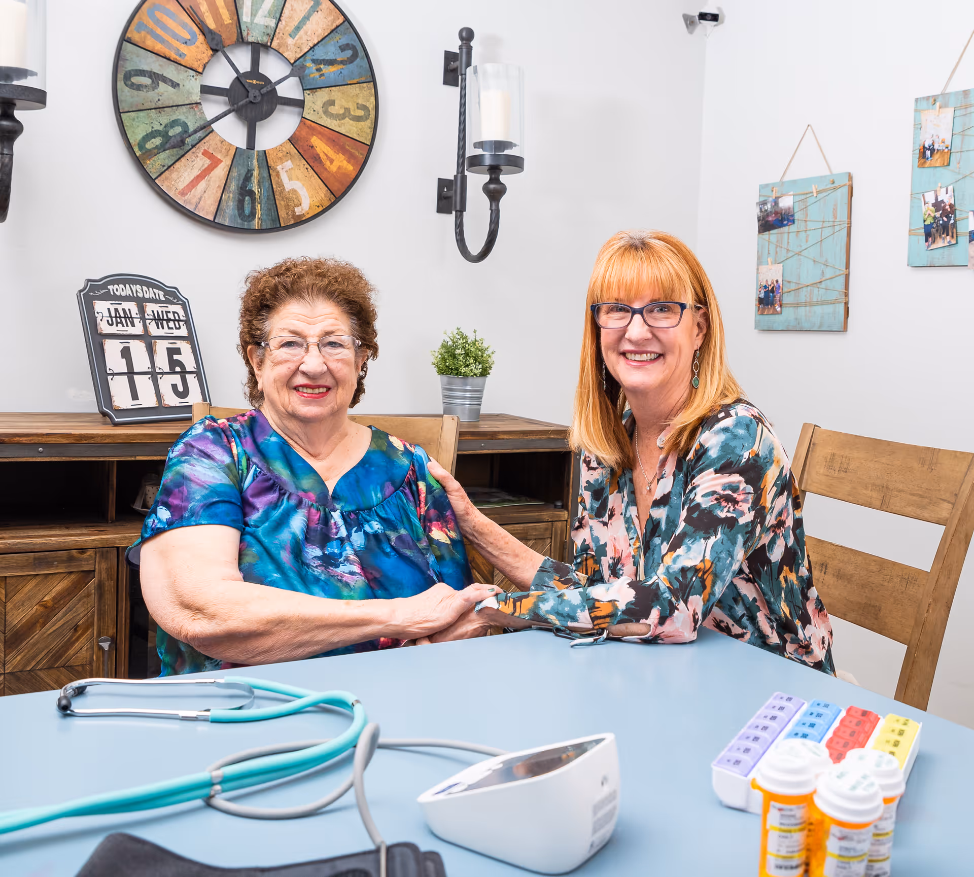 Two women sitting at a table in a cozy room, smiling and holding hands. On the table are a stethoscope, a blood pressure monitor, pill organizers, and prescription bottles. Behind them is a wooden cabinet with a calendar showing January 15, a colorful wall clock, a small potted plant, and wall decorations with photos.