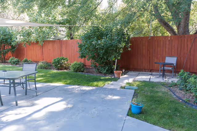 Outdoor patio area with concrete flooring, metal tables and chairs, surrounded by green grass, bushes, trees, and a tall red wooden fence in the background.
