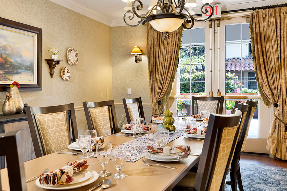 A formal dining room with a long table set for a meal, upholstered chairs, a chandelier, and French doors with curtains leading outside.