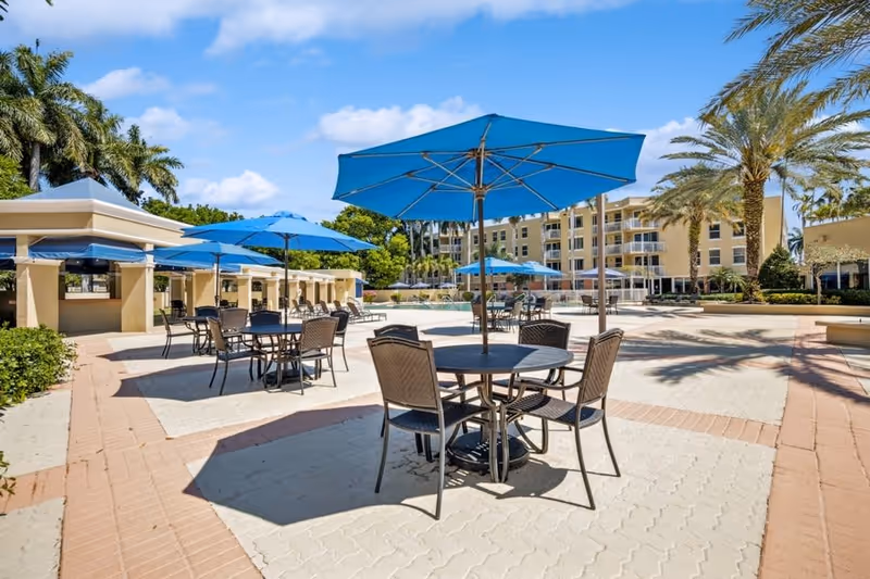 Outdoor patio area with multiple round tables and chairs shaded by blue umbrellas, surrounded by palm trees and a multi-story beige building in the background under a clear blue sky.