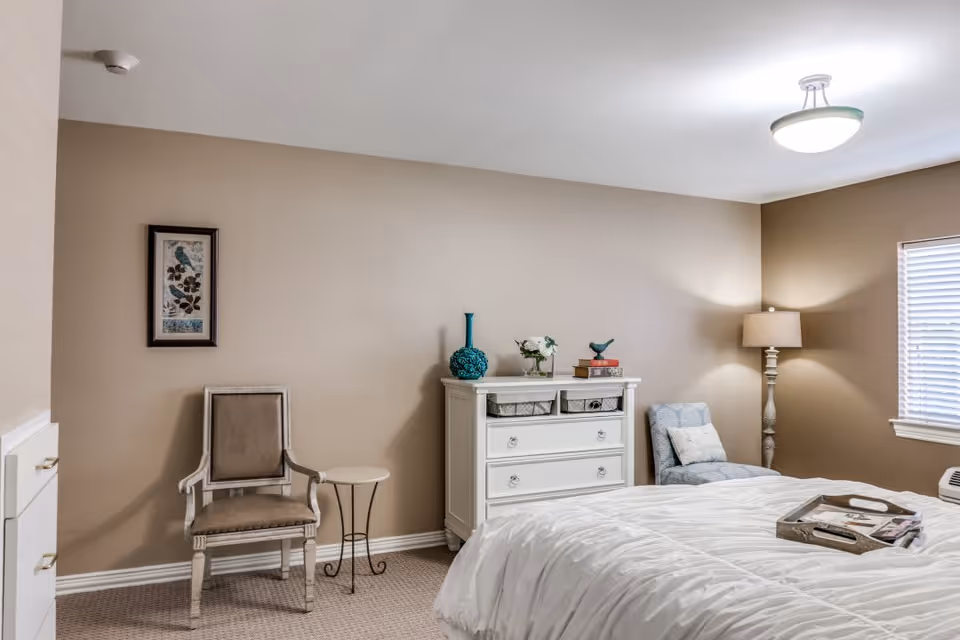 A cozy bedroom with beige walls and carpeted floor. The room features a white dresser with decorative items on top, a beige upholstered chair next to a small round side table, a blue patterned armchair with a cushion, a floor lamp, and a bed with a white comforter and a wooden tray on it. A window with blinds allows natural light into the room.