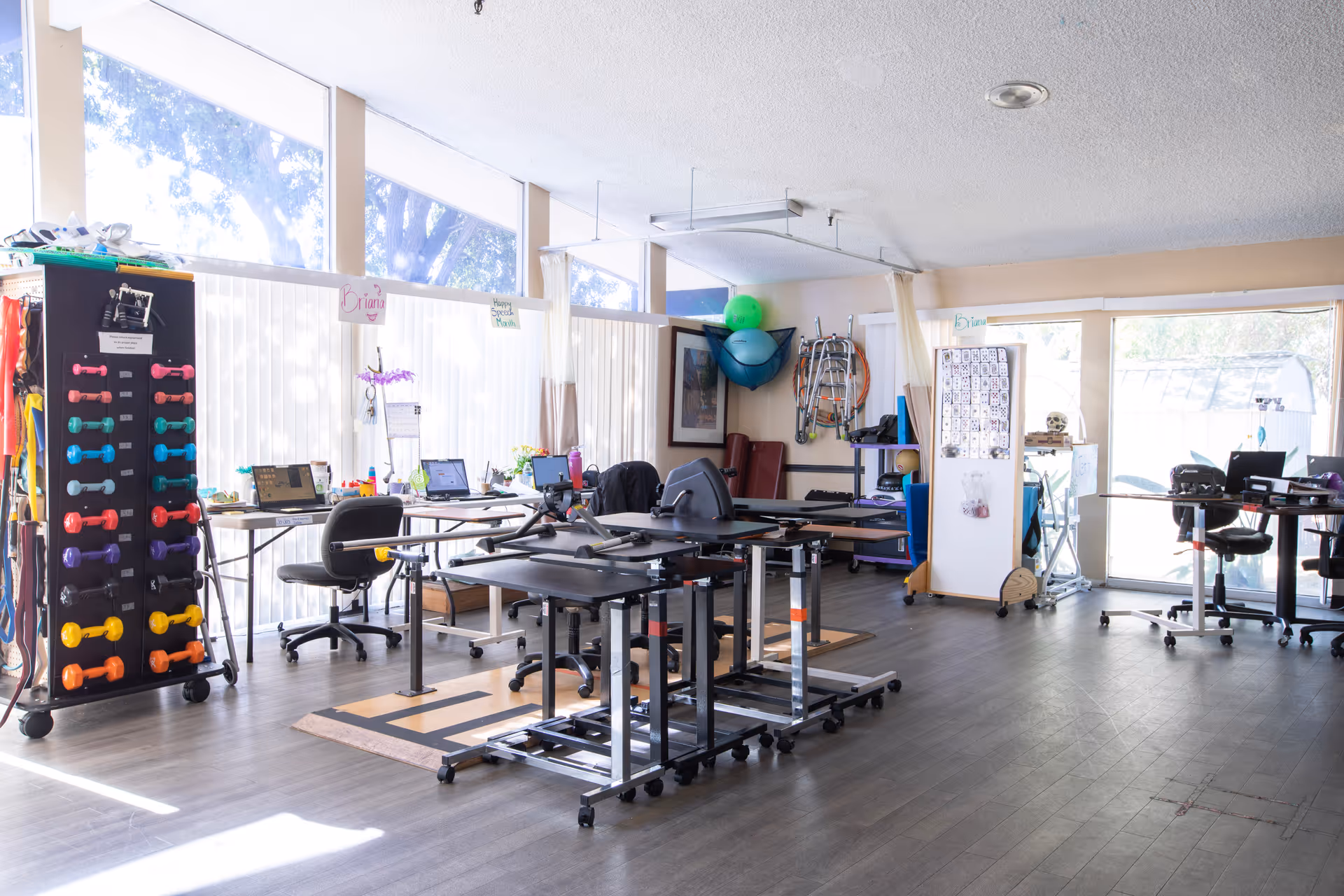A bright room with large windows covered by vertical blinds, featuring exercise equipment including a rack of colorful dumbbells, adjustable tables, office chairs, and various therapy tools. The room appears to be a rehabilitation or therapy area with natural light coming through the windows.