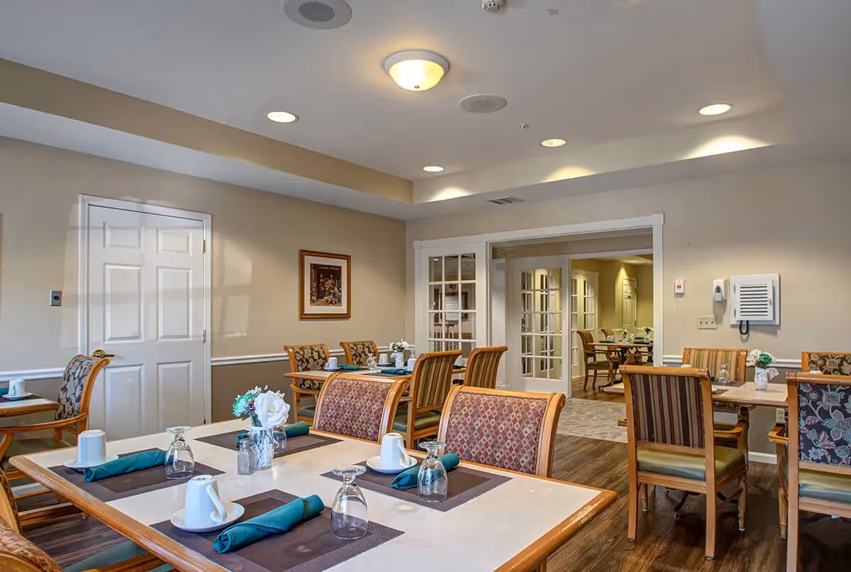 A dining room in a senior living facility with several tables set with white cups, glasses, green napkins, and small flower arrangements. The room has wooden chairs with patterned upholstery, beige walls, recessed lighting, and a door with glass panels leading to another room with more tables and chairs.