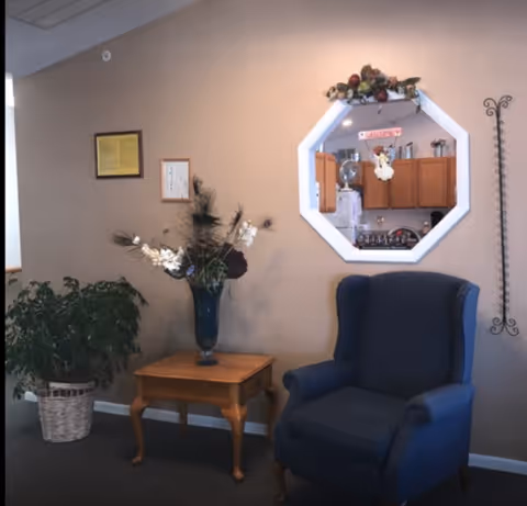 A cozy corner in a living area featuring a blue upholstered armchair, a wooden side table with a decorative vase of flowers, a potted plant in a wicker basket, and a wall-mounted octagonal mirror reflecting a kitchen area with wooden cabinets and appliances. The wall is beige with two framed pictures and a decorative metal wall piece.