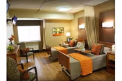 A well-lit senior living facility room with two hospital-style beds, each with orange blankets and pillows. The room has wooden flooring, a window with blinds, a small table with a lamp, chairs, and a wall-mounted TV. The walls are painted in warm tones, and there is a curtain that can be drawn between the beds for privacy.