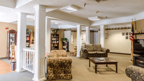 A senior living common area with upholstered sofas, white columns, a coffee table, bookshelf, and a television.