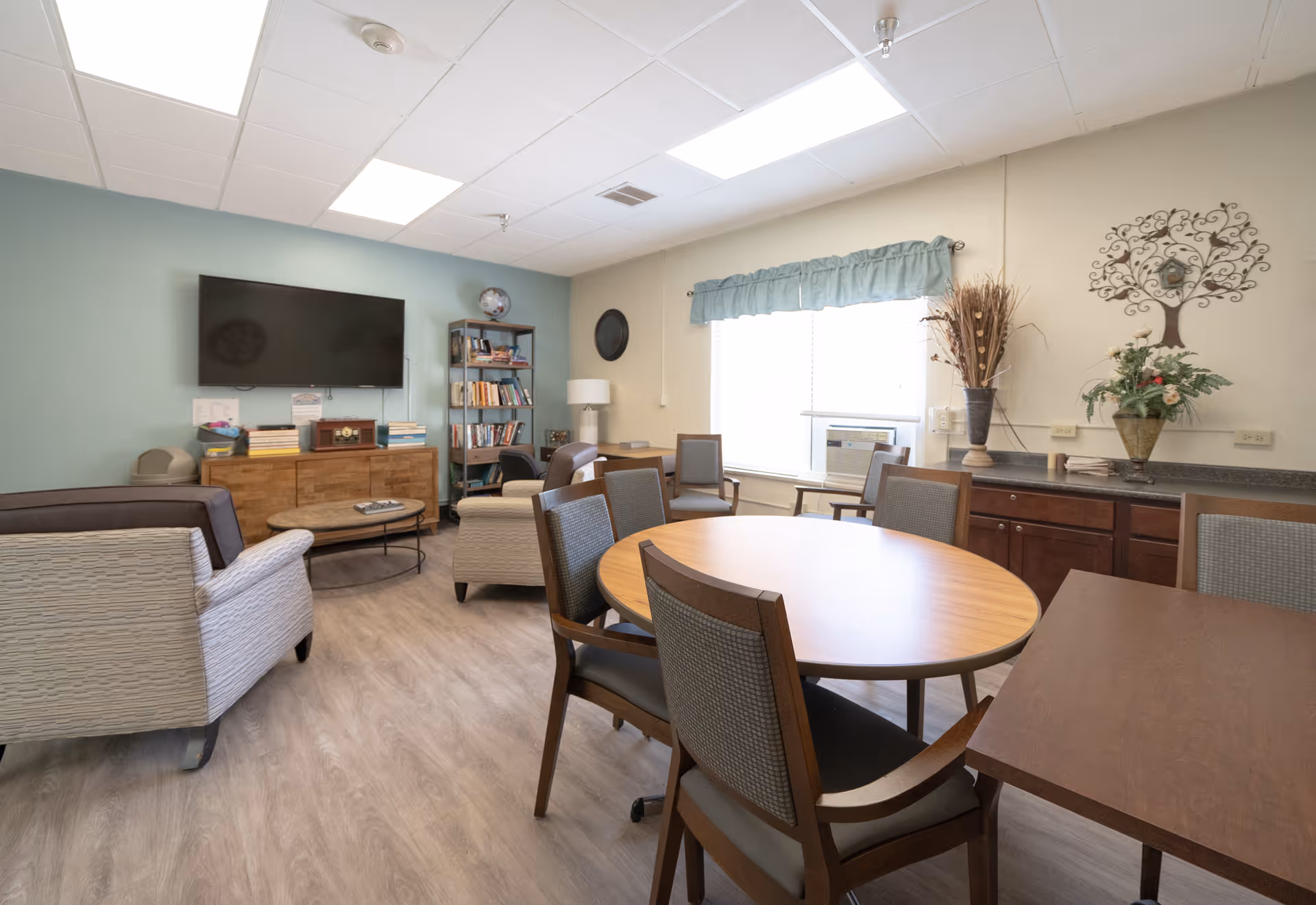 A bright and cozy common area in a senior living facility featuring a round wooden table with chairs, several armchairs, a flat-screen TV mounted on a light blue wall, a bookshelf filled with books, a window with a blue valance, and decorative plants and wall art.