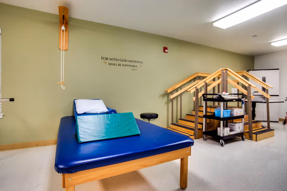 A physical therapy room with a blue padded treatment table, practice stairs and a cart of equipment against a green wall with a motivational quote.
