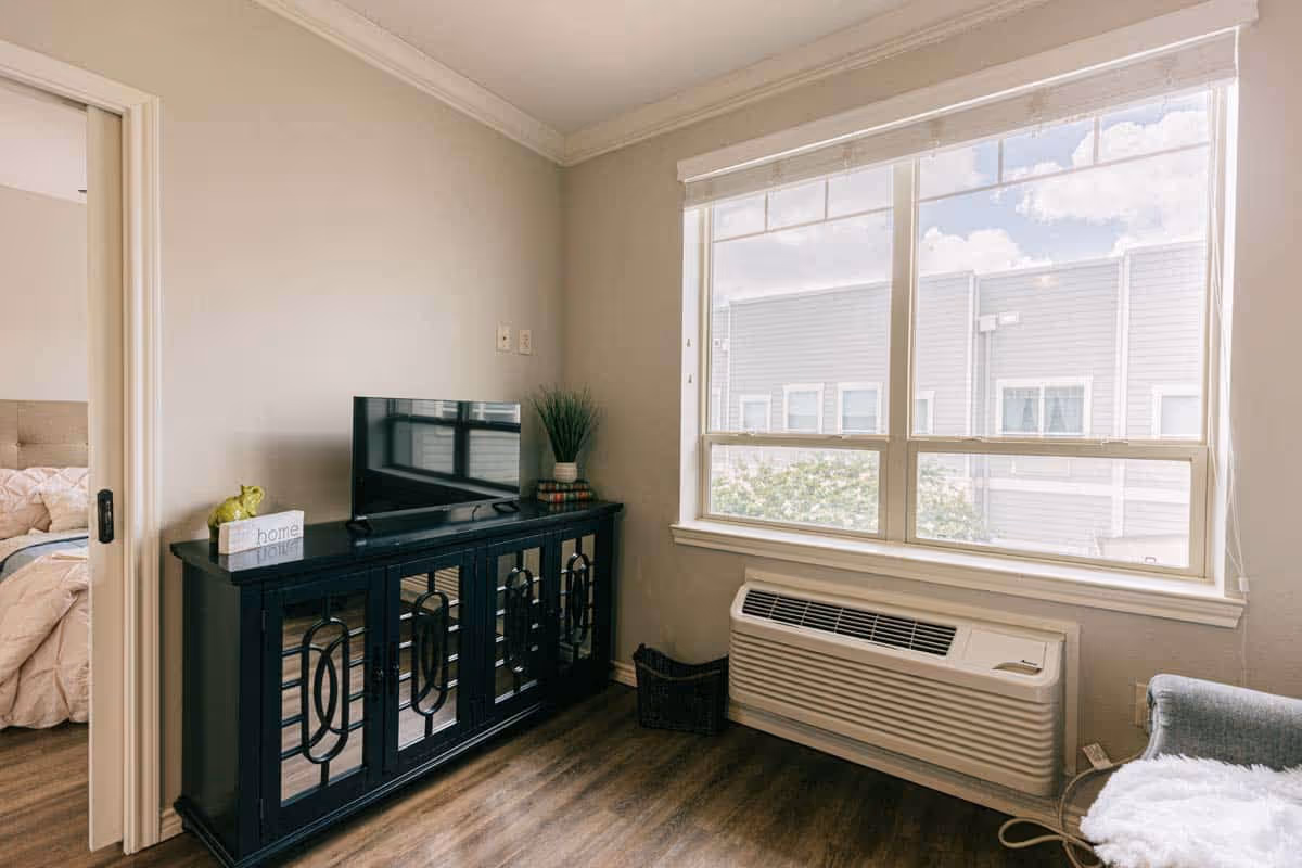 A bright room with a large window showing an outside view of neighboring buildings. Below the window is a wall-mounted air conditioning unit. To the left, there is a black cabinet with a flat-screen TV on top, along with a small decorative plant and a block sign that says 'home'. A doorway on the left leads to a bedroom with a bed visible. The floor is wooden, and a small basket is placed near the air conditioning unit.