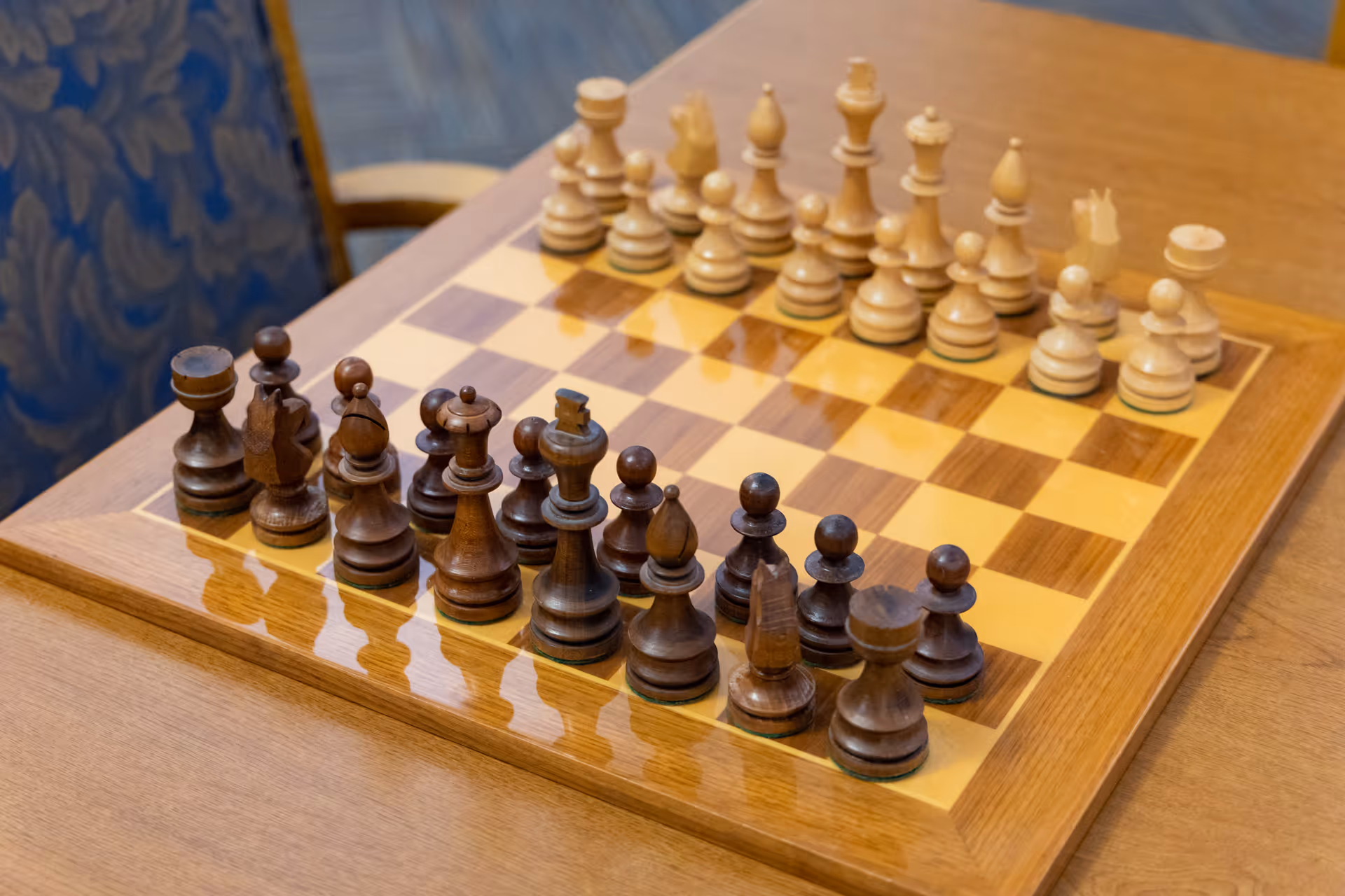 A wooden chessboard with all chess pieces set up in their starting positions on a wooden table, with a blue patterned chair visible in the background.
