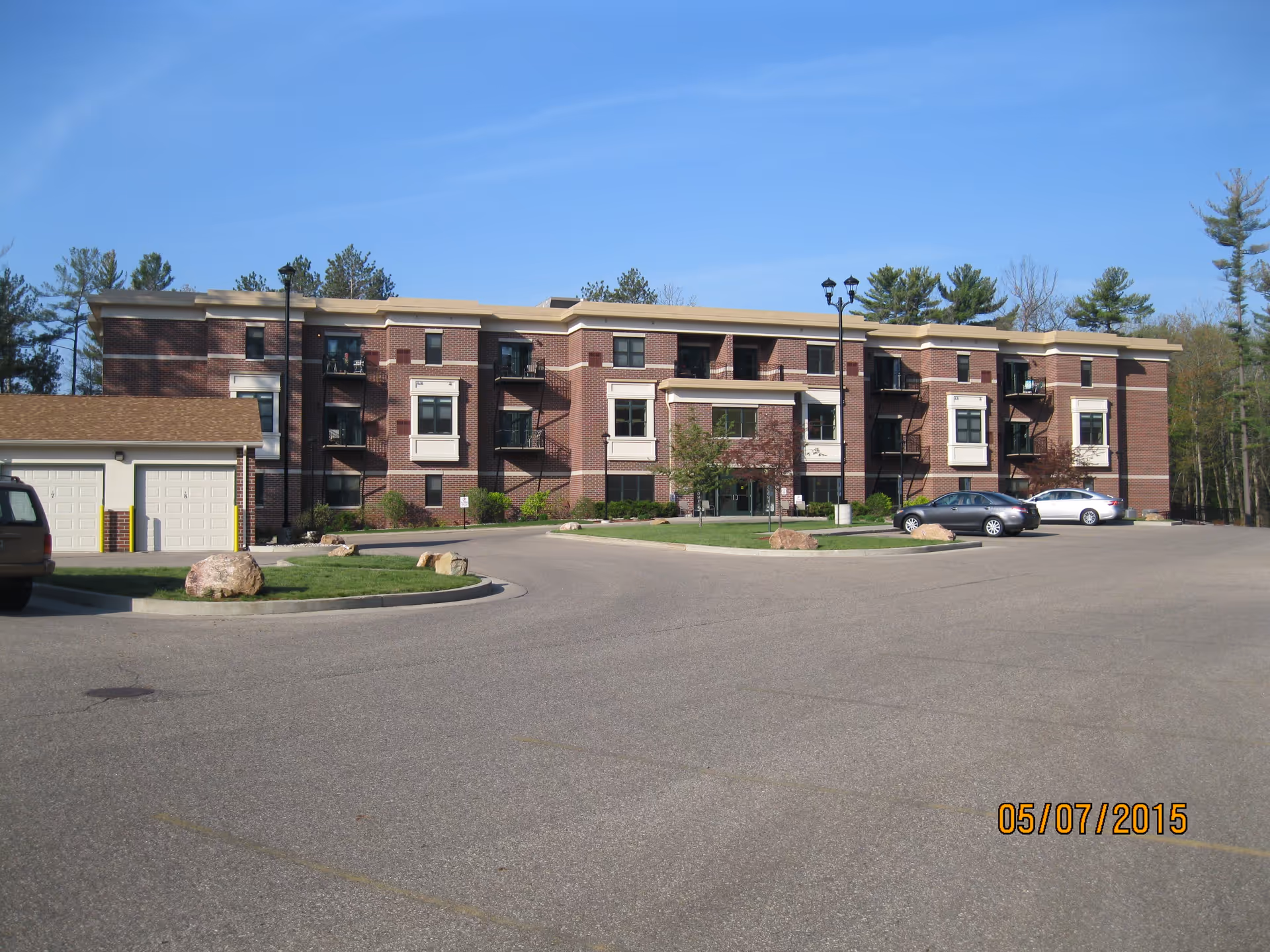 Exterior view of a three-story brick building with multiple windows and small balconies, surrounded by a parking lot with several cars and a small garage structure to the left. Trees are visible in the background under a clear blue sky.