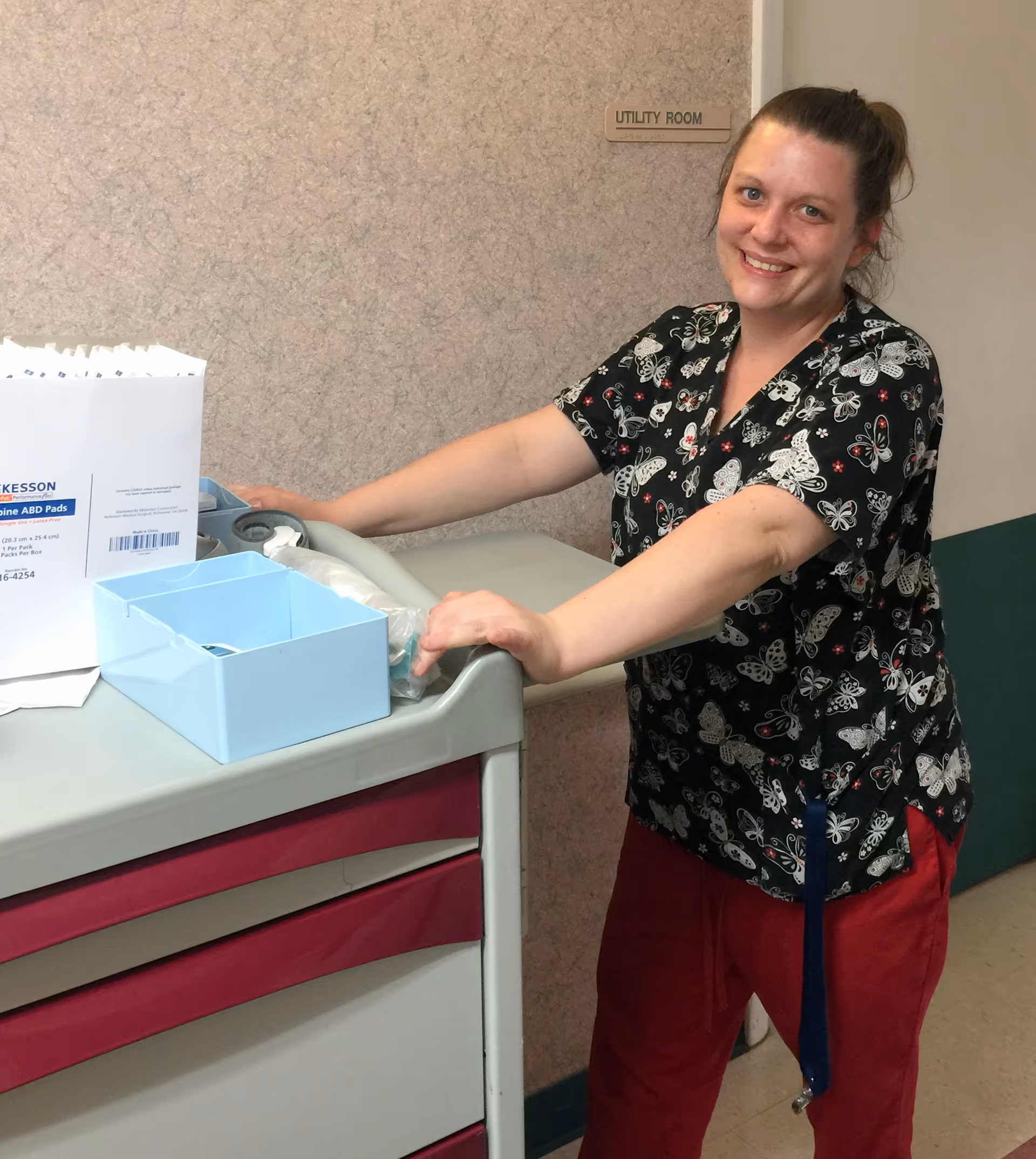A smiling healthcare worker wearing butterfly-patterned scrubs and red pants stands next to a medical cart with supplies in a hallway near a door labeled 'UTILITY ROOM'.