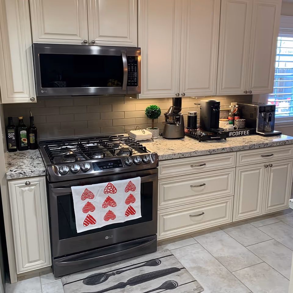 A modern kitchen with white cabinets and granite countertops. There is a stainless steel gas stove with a microwave above it. On the countertop, there are various kitchen appliances including a coffee maker, a juicer, and bottles of olive oil. A white towel with red heart patterns hangs on the oven door. A small decorative plant is also on the counter near the appliances.