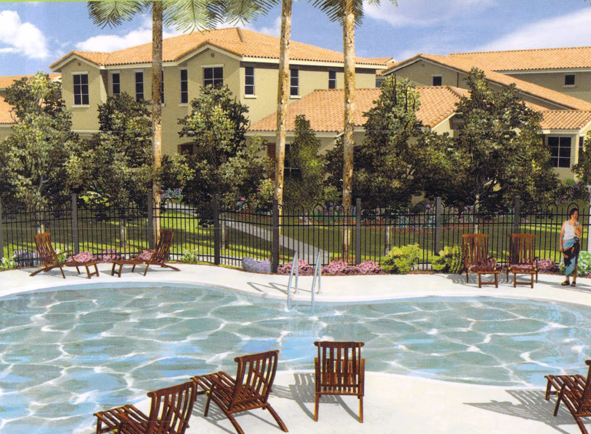 Outdoor swimming pool area with several wooden lounge chairs arranged around the pool. Behind the pool is a black metal fence, trees, and a large building with a tan exterior and red-tiled roof. A person is standing near the fence on the right side of the image.