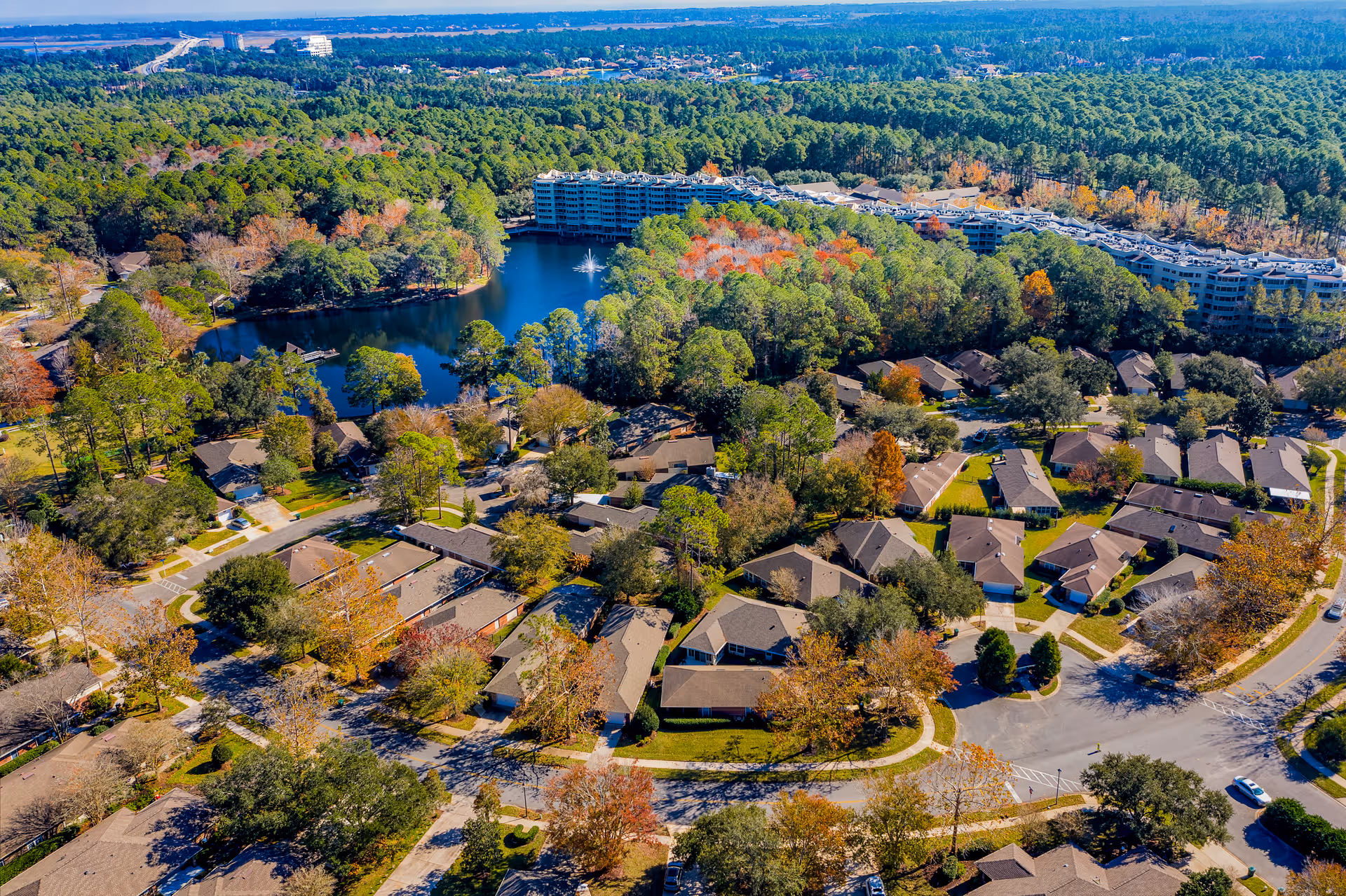 Aerial view of a senior living community with numerous single-story houses surrounded by trees and greenery. In the background, there is a large pond with a fountain and a multi-story building along the water's edge. The area is lush with trees showing autumn colors under a clear sky.