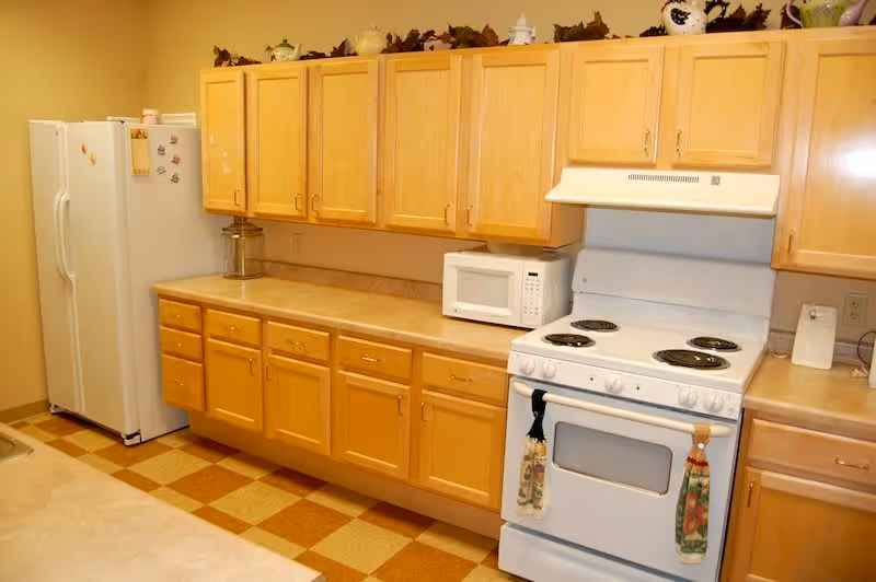A kitchen with light wood cabinets, a white refrigerator, a white microwave, and a white electric stove with two decorative towels hanging on the oven door. The floor has a checkered pattern with beige and brown tiles.